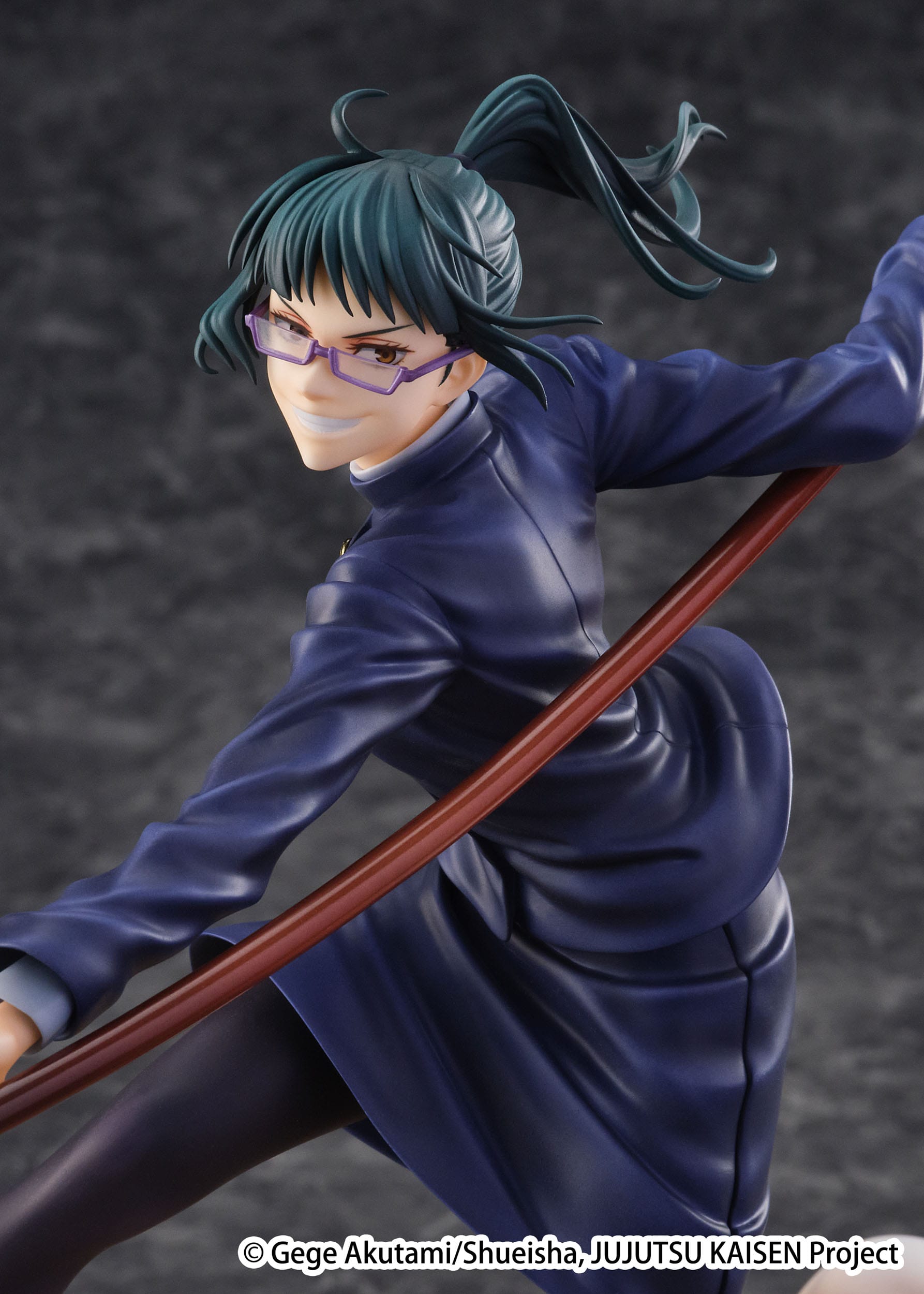 Figurine de Maki Zenin de la série Jujutsu Kaisen en uniforme bleu foncé, en posture dynamique avec un sourire confiant, tenant un long bâton courbé. Vue de face à mi-distance.
