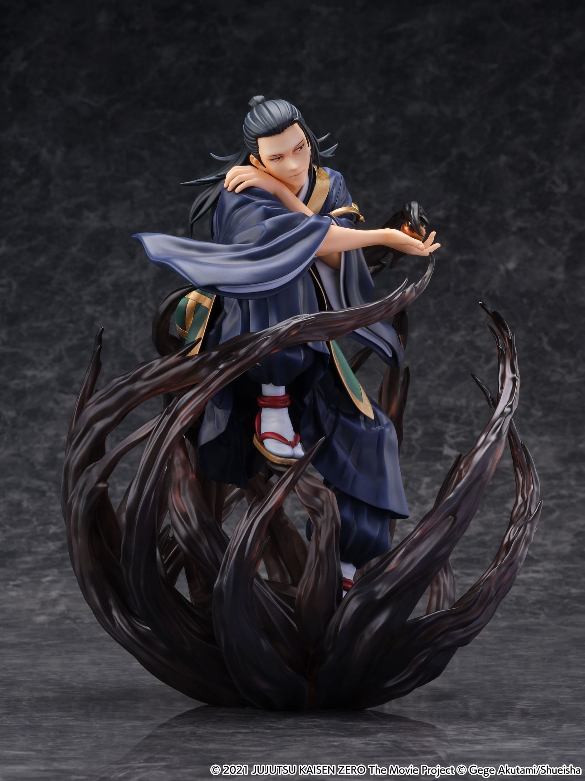 Figurine de Suguru Geto de la série Jujutsu Kaisen Zero en kimono bleu avec accents dorés, en posture dynamique avec expression sérieuse, tenant un objet lumineux. Vue 3/4 à mi-distance.