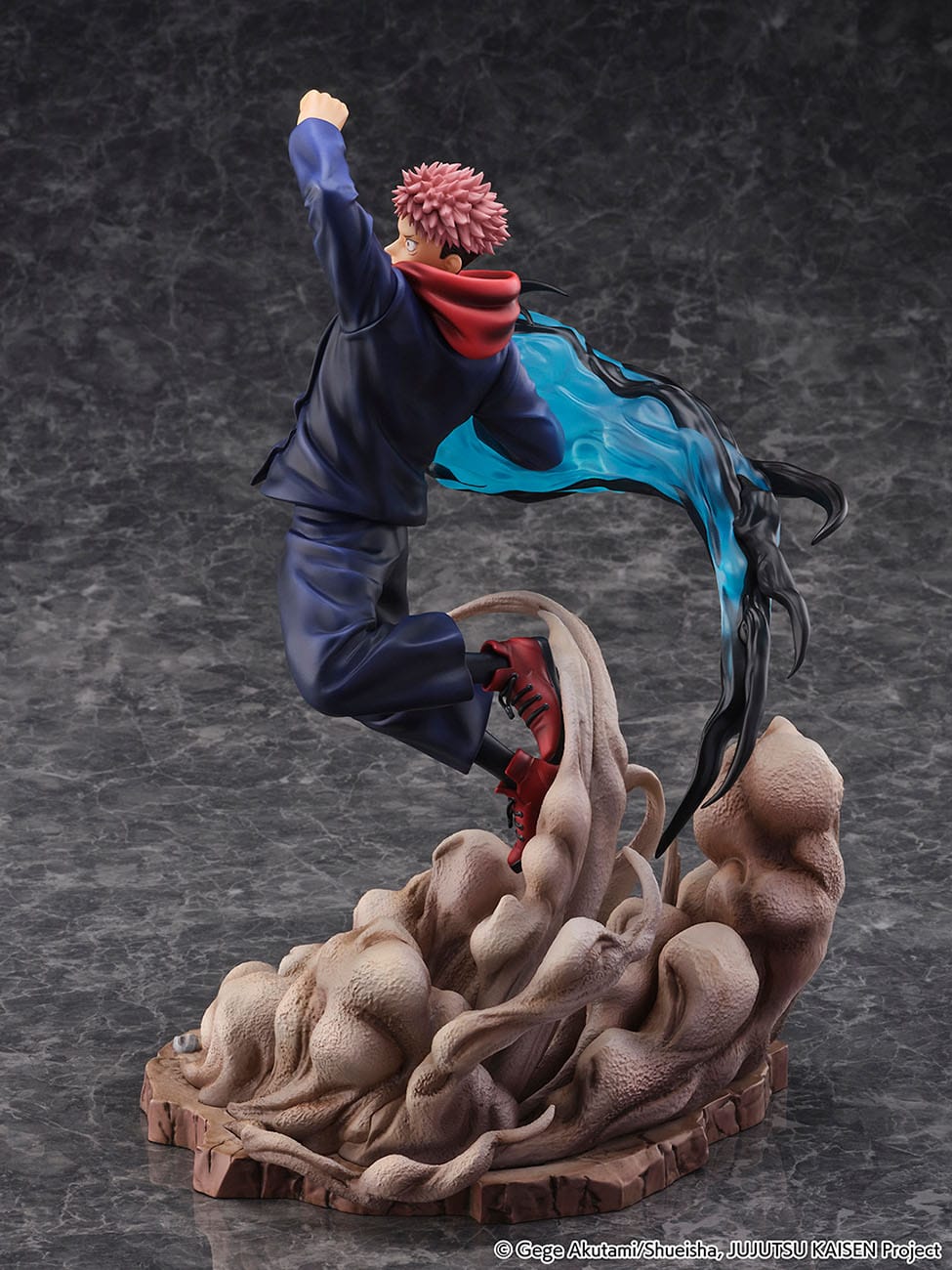 Figurine de Yuji Itadori de la série Jujutsu Kaisen en uniforme bleu foncé avec capuche rouge, en plein saut avec expression déterminée, entouré d'une énergie noire et bleue. Vue de face à mi-distance.