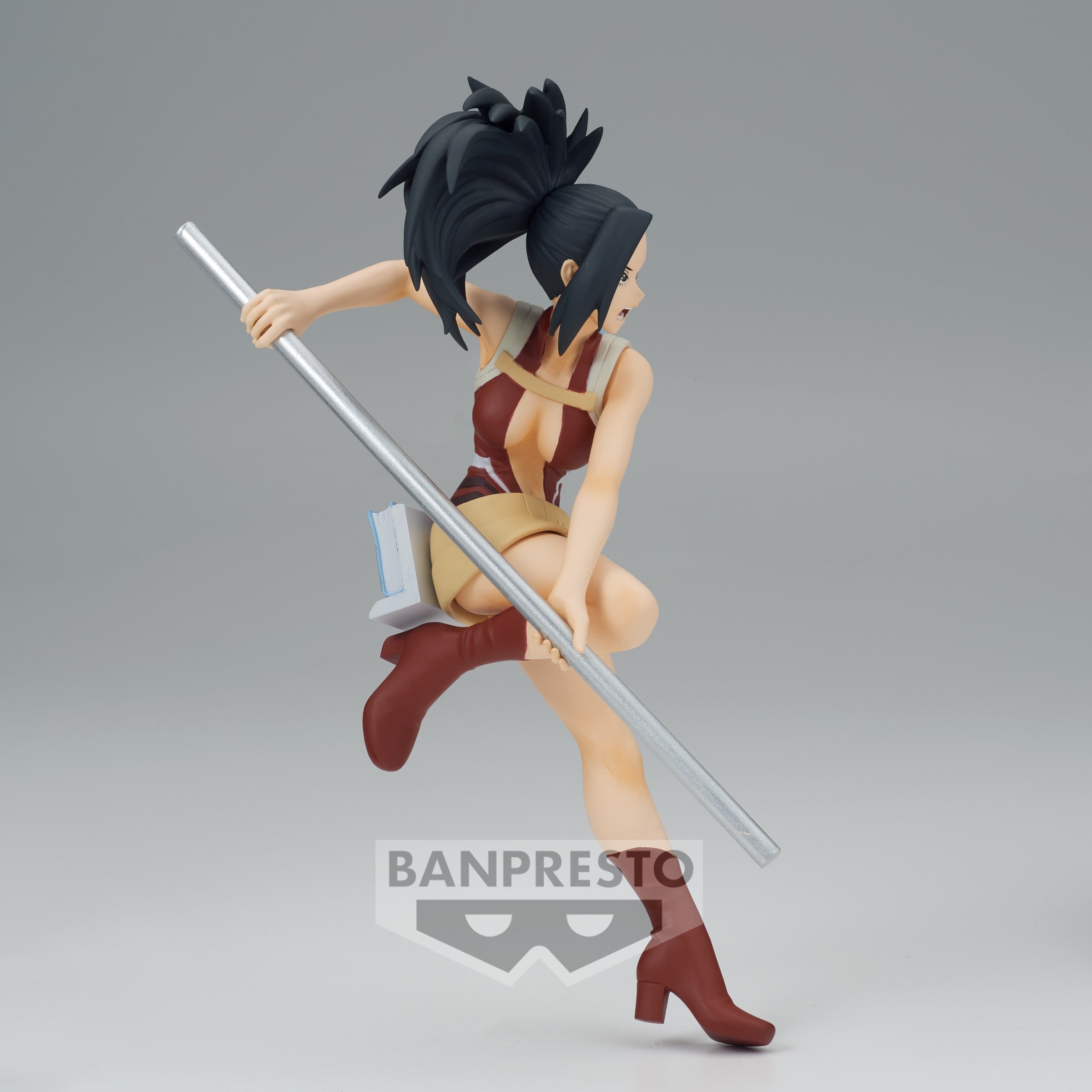 Vue 3/4 arrière à mi-distance de la figurine Momo Yaoyorozu de My Hero Academia en tenue rouge et beige, tenant un bâton métallique.