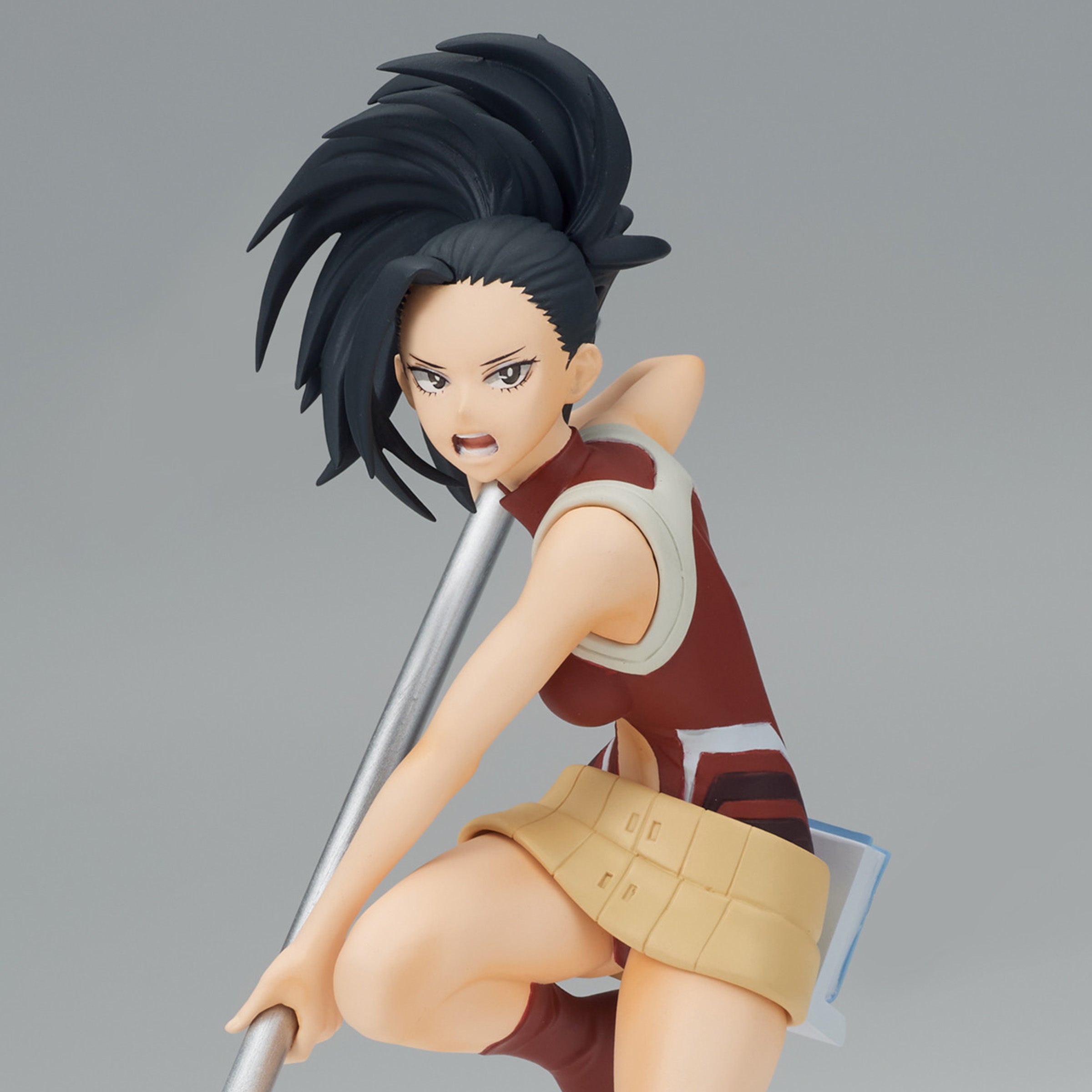 Vue 3/4 de face à mi-distance de la figurine de Momo Yaoyorozu de My Hero Academia en haut sans manches rouge et jupe beige, tenant un bâton.