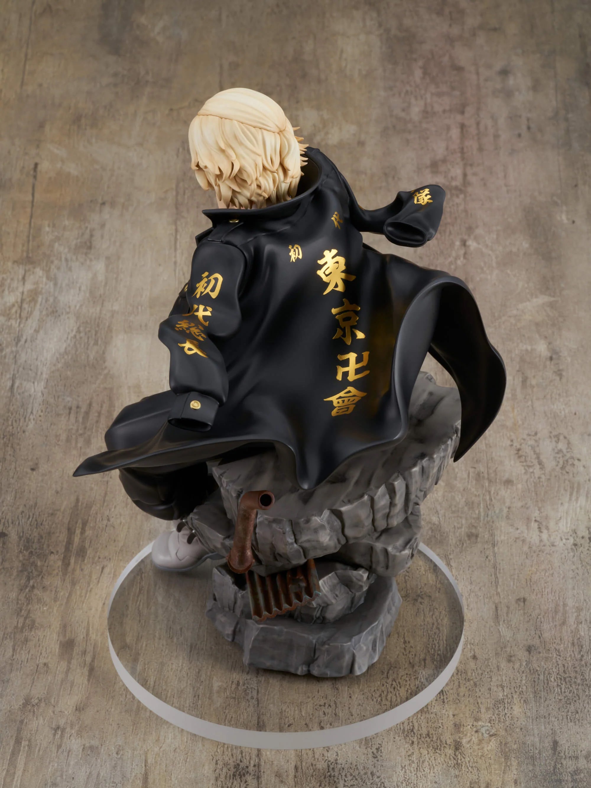 Vue 3/4 arrière à mi-distance de la figurine de Manjiro Sano (Mikey) de Tokyo Revengers en veste noire à lettrage doré, sur un socle rocheux.