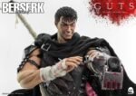 Vue 3/4 de face à mi-distance de la figurine de Guts de Berserk en tenue sombre, tenant une arme à feu tachée de sang.