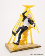Figurine Deluxe de Gojo Satoru de la série Jujutsu Kaisen en veste noire, en posture dynamique avec expression confiante, main pointée vers le haut, entouré d'effets d'énergie jaune. Vue de 3/4 à mi-distance.