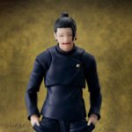 Figurine SH Figuarts de Suguru Geto de la série Jujutsu Kaisen en uniforme bleu foncé, en posture joueuse avec un sourire malicieux, vue de face à mi-distance.